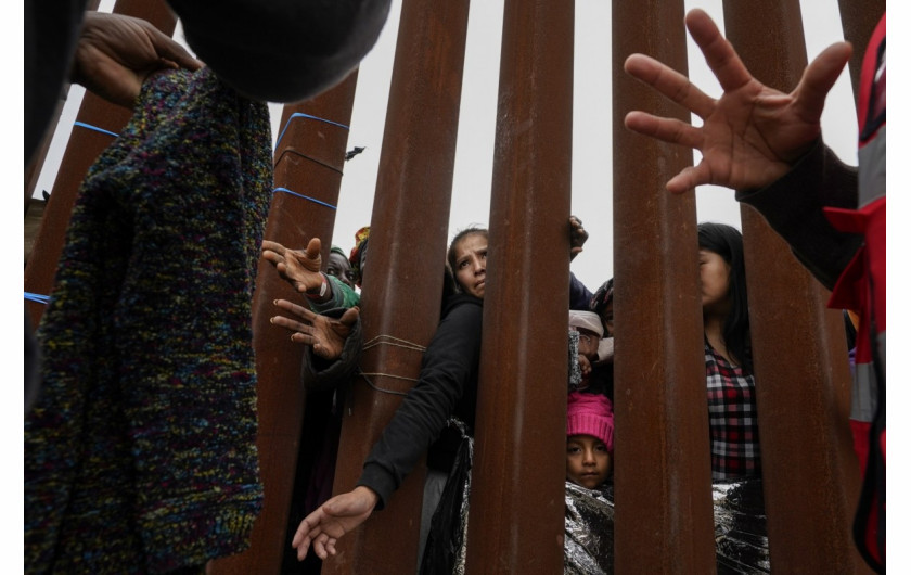 Migranci sięgają przez mur graniczny po odzież rozdawaną przez wolontariuszy, gdy czekają między dwoma murami granicznymi, aby ubiegać się o azyl w piątek, 12 maja 2023 r., w San Diego. Setki migrantów czekają między dwoma murami, wielu z nich przez wiele dni. (AP Photo/Gregory Bull)