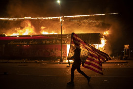 Protestujący niesie amerykańską flagę do góry nogami. Znak niepokoju obok płonącego budynku, 28 maja 2020 r. w Minneapolis. Trzecia noc protestów w Minneapolis, związanych ze śmiercią George'a Floyda (Zdjęcie: Julio Cortez.) / Pulitzer Prize 2021 for Breaking News Photography