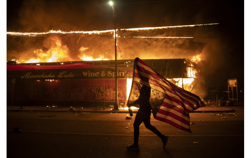 Protestujący niesie amerykańską flagę do góry nogami. Znak niepokoju obok płonącego budynku, 28 maja 2020 r. w Minneapolis. Trzecia noc protestów w Minneapolis, związanych ze śmiercią George'a Floyda (Zdjęcie: Julio Cortez.) / Pulitzer Prize 2021 for Breaking News Photography