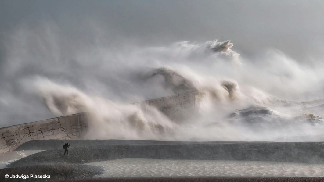 &lt;strong&gt;II miejsce, fot. Jadwiga Piasecka, Wielka Brytania&lt;/strong&gt;&lt;br /&gt;&lt;br /&gt;

„Od dziecka fascynowały mnie wielkie fale i sztormy – potęga i energia morza zawsze robiły na mnie ogromne wrażenie. Gdy więc nadciągnął sztorm Eunice, wiedziałam, że nie mogę przegapić okazji, by zobaczyć go na własne oczy” - mówi fotografka Jadwiga Piasecka.&lt;br /&gt;&lt;br /&gt;

Zdjęcie wykonała z bezpiecznego, osłoniętego miejsca w Newhaven, na południowym wybrzeżu Wielkiej Brytanii, gdzie porywy wiatru przekraczały 80 mil na godzinę. „Z mojej perspektywy obserwowałam, jak ogromne fale uderzają w falochron, wyrzucając w powietrze dramatyczne kaskady wody... To naprawdę pokazywało skalę i siłę żywiołu” - wspomina.&lt;br /&gt;&lt;br /&gt;

Sztorm Eunice przyniósł najwyższy zarejestrowany w Anglii podmuch wiatru - 122 mile na godzinę. Był jednym z kilku gwałtownych niżów europejskich na początku 2022 roku, które spowodowały poważne zniszczenia w Wielkiej Brytanii i Irlandii: rekordowe wichury, uszkodzenia infrastruktury oraz zalania przybrzeżne. Tak potężne fale powstają w wyniku połączenia silnego wiatru z niskim ciśnieniem w centrum niżu, co powoduje spiętrzanie się morza w wysokie grzywacze. Gdy uderzają one w nadbrzeżne konstrukcje, wyrzucają w powietrze ogromne ilości wody.&lt;br /&gt;&lt;br /&gt;

Jeden z jurorów konkursu, Peter Gibbs, zauważył: „Sylwetka samotnej postaci podkreśla potęgę i skalę sił natury, a zmarszczki na wodzie na pierwszym planie dodatkowo akcentują siłę wiatru.” Na szczęście większość osób zastosowała się wówczas do rzadko wydawanego czerwonego ostrzeżenia pogodowego Met Office i nie zbliżała się do miejsca, gdzie szalał sztorm.
