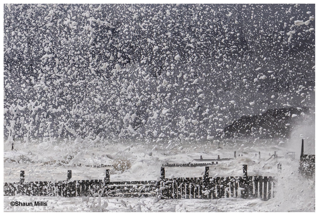 &lt;strong&gt;III Nagroda publiczności, fot. Shaun Mills, Wielka Brytania&lt;/strong&gt;&lt;br /&gt;&lt;br /&gt;

Podczas sztormu na wschodnim wybrzeżu Wielkiej Brytanii fale uderzają o nadbrzeżne umocnienia w Overstrand w hrabstwie Norfolk, wyrzucając słupy morskiej piany wysoko w powietrze. Fotograf Shaun Mills wspomina: „Podczas fotograficznego wyjazdu na wybrzeże Norfolk trafiłem na sztorm i postanowiłem wykorzystać okazję, by uchwycić niezwykle wzburzone morze. Zrobiłem to zdjęcie, aby wyizolować pojedyncze rozpryski wody, nadając całości wrażenie śnieżnej zamieci”.&lt;br /&gt;&lt;br /&gt;

Morska mgiełka powstaje, gdy silny wiatr rozrywa grzbiety fal, wyrzucając w powietrze drobne kropelki słonej wody. Te unoszące się cząsteczki mogą mieć różną wielkość - od widocznych rozprysków po mikroskopijne drobiny, które wpływają na powstawanie chmur i procesy pogodowe. Uwiecznienie ich w ruchu wymaga krótkiego czasu naświetlania i precyzyjnego wyczucia momentu. W tym przypadku kadr został przycięty tak, aby wypełnić całą przestrzeń dynamiczną fakturą rozpryskującej się wody.&lt;br /&gt;&lt;br /&gt;

Burzowe warunki, które tworzą takie sceny, stają się coraz częstsze i bardziej intensywne w miarę ocieplania się klimatu. Wraz ze wzrostem globalnych temperatur rośnie energia zasilająca ekstremalne zjawiska pogodowe. Jednocześnie podnoszący się poziom mórz stanowi coraz większe zagrożenie dla nisko położonych obszarów przybrzeżnych, takich jak Norfolk. Wybrzeże East Anglii jest szczególnie podatne na erozję z powodu miękkich, kruchych klifów i osiadającego gruntu. Połączenie silnych sztormów z wyższym poziomem morza przyspiesza erozję brzegową i stanowi coraz poważniejsze wyzwanie dla przestarzałych umocnień nadmorskich.
