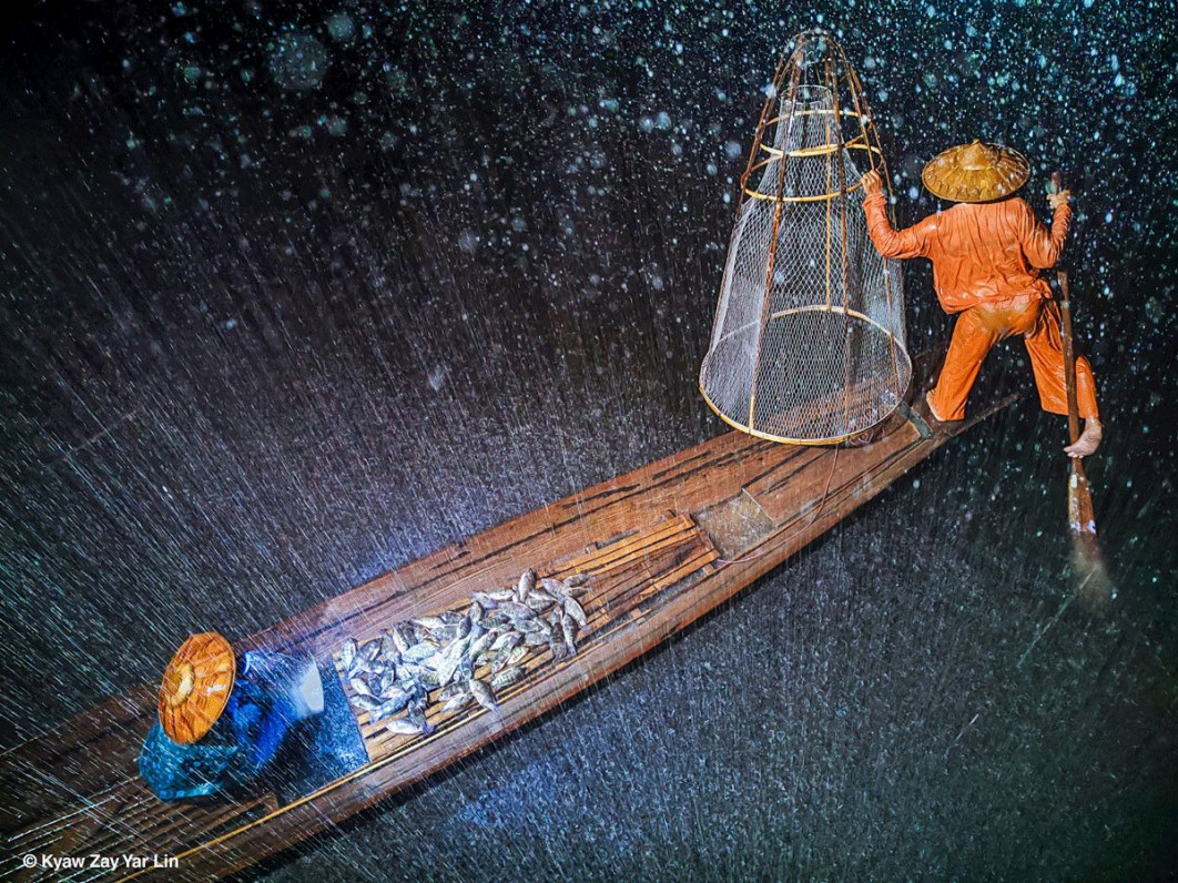 &lt;strong&gt;I miejsce w kat. Fotografia mobilna, fot. Kyaw Zay Yar Lin,  Mjanma&lt;/strong&gt;&lt;br /&gt;&lt;br /&gt;

Zdjęcie oddaje napięcie chwili, gdy tropikalny deszcz spada nagle i gwałtownie. Dwóch rybaków działa w pośpiechu - jeden wiosłuje po ciemnej wodzie, drugi wylewa wodę z dna łodzi. Ich jaskrawe pomarańczowe i niebieskie ubrania kontrastują z szarością otoczenia, a smugi deszczu i rozmazany ruch postaci sprawiają, że widz ma wrażenie, jakby sam znalazł się w środku ulewy. Krople spadają gęsto, rozpryskując się na powierzchni jeziora, podczas gdy łódź stopniowo wypełnia się wodą.&lt;br /&gt;&lt;br /&gt;

Wioślarz korzysta z charakterystycznej dla ludu Intha techniki - stoi na rufie, owijając jedną nogę wokół wiosła. Pozwala to na płynne i precyzyjne manewrowanie między trzcinami jeziora. Choć Inle Lake jest płytkie - jego średnia głębokość nie przekracza dwóch metrów – od wieków stanowi centrum lokalnego ekosystemu i podstawę życia mieszkańców. Dziś jednak ten sposób życia znajduje się w coraz większym zagrożeniu.&lt;br /&gt;&lt;br /&gt;

Monsunowy klimat Mjanmy przynosi intensywne opady, a rybołówstwo pozostaje niezbędnym źródłem utrzymania, nawet podczas burz. Jednak wylesianie okolicznych wzgórz powoduje erozję gleby - osady spływają do jeziora, które skurczyło się już do połowy swojej pierwotnej powierzchni. Dodatkowo zmiany klimatyczne zaburzają rytm opadów i przyspieszają parowanie, przez co poziom wody w jeziorze waha się coraz bardziej. W przypadku tak płytkiego zbiornika zjawiska te zagrażają zarówno populacjom ryb, jak i delikatnemu ekosystemowi, który od pokoleń kształtuje życie nad Inle Lake.
