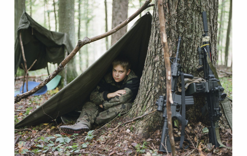 fot. Natalia Kepesz, Polska, z cyklu: Niewybuch, 3 miejsce w kategorii Portret - seria zdjęć / World Press Photo 2021


Wojskowe obozy letnie dla młodzieży istnieją w Polsce od lat 20. ubiegłego wieku. Młodzi uczestnicy przechodzą przez obozy kondycyjne, poddawani są ciężkim próbom fizycznym i psychicznym, a także uczą się - często na byłych poligonach wojskowych - taktyki, przetrwania, samoobrony i topografii. Uczy się ich także strzelania, używając karabinów pneumatycznych, a czasem replik broni, takich jak karabiny maszynowe i granatniki. Obozy promowane są jako okazja do przeżycia przygody i rekreacji, a także jako zajęcia kształtujące charakter i zachęcające do pracy w grupie. Organizatorzy utrzymują, że uczestnictwo w grach z replikami broni powstrzymuje dzieci przed sięgnięciem po prawdziwą. Z drugiej strony, pojawiają się głosy krytyki, które sugerują, że popularność obozów wynika ze wzrostu nacjonalizmu w Polsce, szczególnie od czasu dojścia do władzy PiS, prawicowej partii populistycznej. Patriotyzm i nacjonalizm odgrywają istotną rolę w edukacji szkolnej.