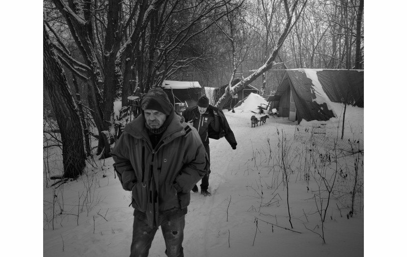 Fot. Maciej Grzybowski, 1. miejsce w kat. Documentary Project / Grand Press Photo 2024Ela, Darek i Mirek są w bezdomności od wielu lat. Mieszkają w lesie nad rzeką, na obrzeżach miasta. Opiekują się dwoma psami Florą i Jogim. Ela przez kilkanaście lat pracowała w zakładach farmaceutycznych. Jest wdową, jej syn mieszka za granicą. Obecnie bezrobotna. Darek, partner Eli, podejmuje się prac dorywczych na budowach i w firmach sprzątających miasto. Sam zbudował wszystkie domki. Mirek pochodzi z Pomorza, jest wolnym duchem. Często wychodzi w nocy zbierać złom, który potem sprzedaje.
W Polsce około 30 tysięcy osób znajduje się w kryzysie bezdomności, z czego ponad 80 proc. stanowią mężczyźni.
