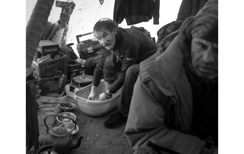 Fot. Maciej Grzybowski, 1. miejsce w kat. Documentary Project / Grand Press Photo 2024Ela, Darek i Mirek są w bezdomności od wielu lat. Mieszkają w lesie nad rzeką, na obrzeżach miasta. Opiekują się dwoma psami Florą i Jogim. Ela przez kilkanaście lat pracowała w zakładach farmaceutycznych. Jest wdową, jej syn mieszka za granicą. Obecnie bezrobotna. Darek, partner Eli, podejmuje się prac dorywczych na budowach i w firmach sprzątających miasto. Sam zbudował wszystkie domki. Mirek pochodzi z Pomorza, jest wolnym duchem. Często wychodzi w nocy zbierać złom, który potem sprzedaje.
W Polsce około 30 tysięcy osób znajduje się w kryzysie bezdomności, z czego ponad 80 proc. stanowią mężczyźni.
