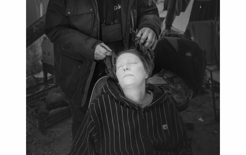Fot. Maciej Grzybowski, 1. miejsce w kat. Documentary Project / Grand Press Photo 2024Ela, Darek i Mirek są w bezdomności od wielu lat. Mieszkają w lesie nad rzeką, na obrzeżach miasta. Opiekują się dwoma psami Florą i Jogim. Ela przez kilkanaście lat pracowała w zakładach farmaceutycznych. Jest wdową, jej syn mieszka za granicą. Obecnie bezrobotna. Darek, partner Eli, podejmuje się prac dorywczych na budowach i w firmach sprzątających miasto. Sam zbudował wszystkie domki. Mirek pochodzi z Pomorza, jest wolnym duchem. Często wychodzi w nocy zbierać złom, który potem sprzedaje.
W Polsce około 30 tysięcy osób znajduje się w kryzysie bezdomności, z czego ponad 80 proc. stanowią mężczyźni.
