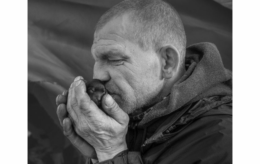 Fot. Maciej Grzybowski, 1. miejsce w kat. Documentary Project / Grand Press Photo 2024Ela, Darek i Mirek są w bezdomności od wielu lat. Mieszkają w lesie nad rzeką, na obrzeżach miasta. Opiekują się dwoma psami Florą i Jogim. Ela przez kilkanaście lat pracowała w zakładach farmaceutycznych. Jest wdową, jej syn mieszka za granicą. Obecnie bezrobotna. Darek, partner Eli, podejmuje się prac dorywczych na budowach i w firmach sprzątających miasto. Sam zbudował wszystkie domki. Mirek pochodzi z Pomorza, jest wolnym duchem. Często wychodzi w nocy zbierać złom, który potem sprzedaje.
W Polsce około 30 tysięcy osób znajduje się w kryzysie bezdomności, z czego ponad 80 proc. stanowią mężczyźni.
