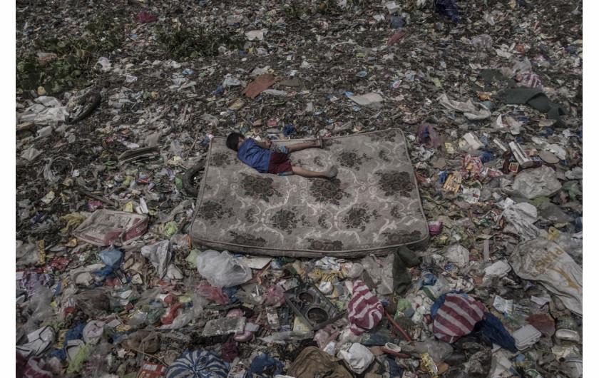 fot. Mario Cruz, Living Among What's Left Behind, 3. miejsce w kategorii Environment.

Dziecko na materacu zbiera produkty nadające się do przetworzenia ze śmieci unoszących się na rzece Pasig na Filipinach. Rzeka ta została ogłoszona biologicznie martwą już w latach 90. XX wieku, w skutek zanieczyszczenia przemysłowego i wypuszczania nieoczyszczonych ścieków przez zamieszkujące jej wybrzeża społeczności. Zaliczana jest do 20 najbardziej zanieczyszczonych rzek na świecie. Co roku z jej nurtem do oceanu wpada 64 tys. ton plastiku. Podejmowane są działania mające za zadanie jej oczyszczenie, ale nadal na niektórych odcinkach zanieczyszczenie jest tak ogromne, że możliwe jest chodzenie po unoszących się na niej odpadach.