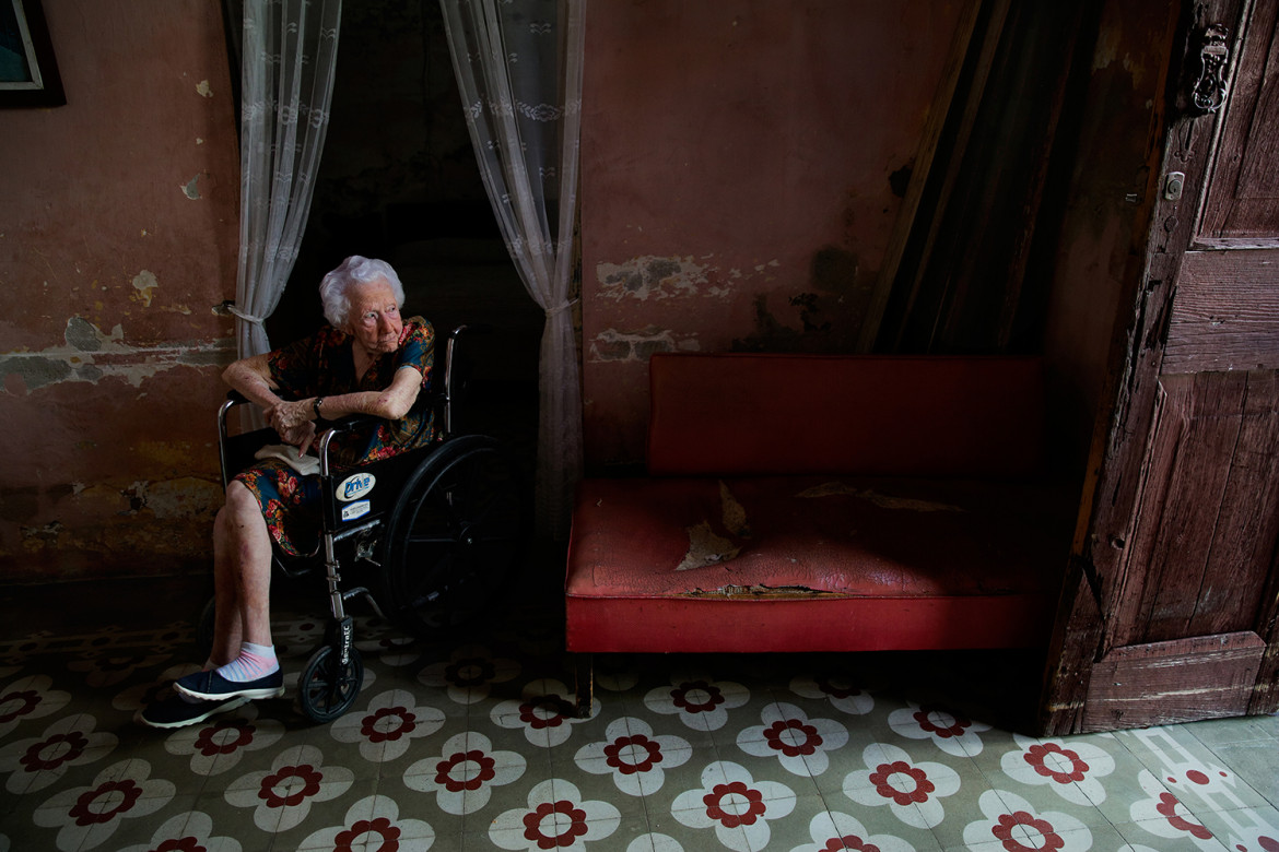 fot. Oded Wagenstein, Onelia Katyo patrząca na ulicę przez drzwi mieszkania, Cienfuegos, Kuba, Marzec 2015. Ze względu na brak światła i klimatyzacji, większość drzwi w małej społeczności pozostaje otwarta przez cały dzień, a każdy kto przechodzi przez drzwi Onelii jest jej przyjacielem: sąsiadem, dziećmi w drodze do pobliskiej szkoły, a nawet bezpańskim kotem. Od 2014 roku spotkaliśmy się siedem razy i nigdy nie widziałem jej w sukience bez motywu kwiatów. Powiedziała mi, że kwiaty sprawiają, że czuje się szczęśliwa.